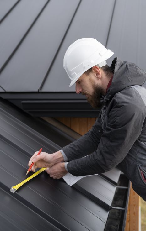 A construction worker wearing a white hard hat measures and marks lines on a black metal roof using a yellow tape measure and red pencil, showcasing the precision involved in installing energy efficient metal roofs.
